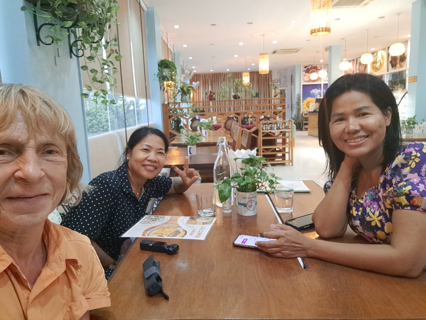 Kolme naista, yksi suomalainen ja kaksi kambodzalaista, hymyilevät yhteiskuvassa kahvilan pöydän ääressä.